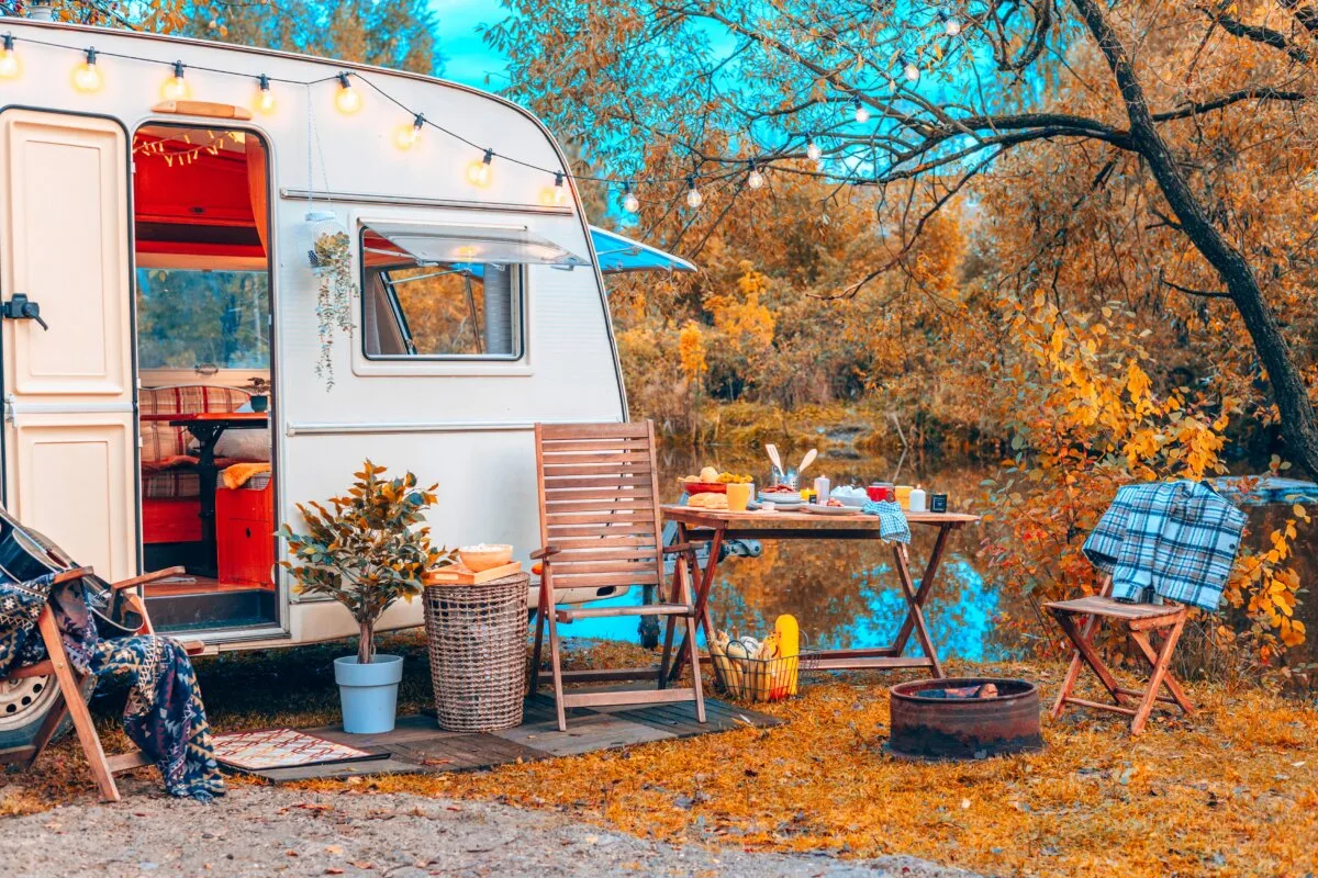 Trailer of mobile home or recreational vehicle stands on shore of pond in camping in autumn near table set