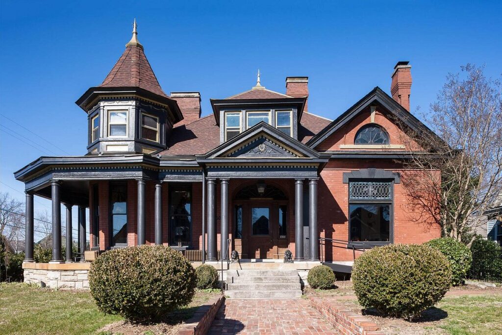 Urban Cowboy Nashville Victorian-style red brick house with ornate trim, turret, and welcoming porch surrounded by lush landscaping.