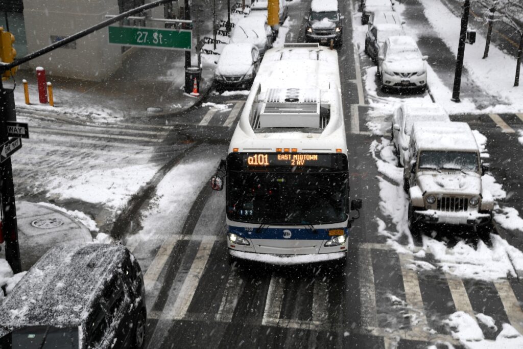 Public transit bus Q101 drives through a snowy urban intersection in East Midtown.