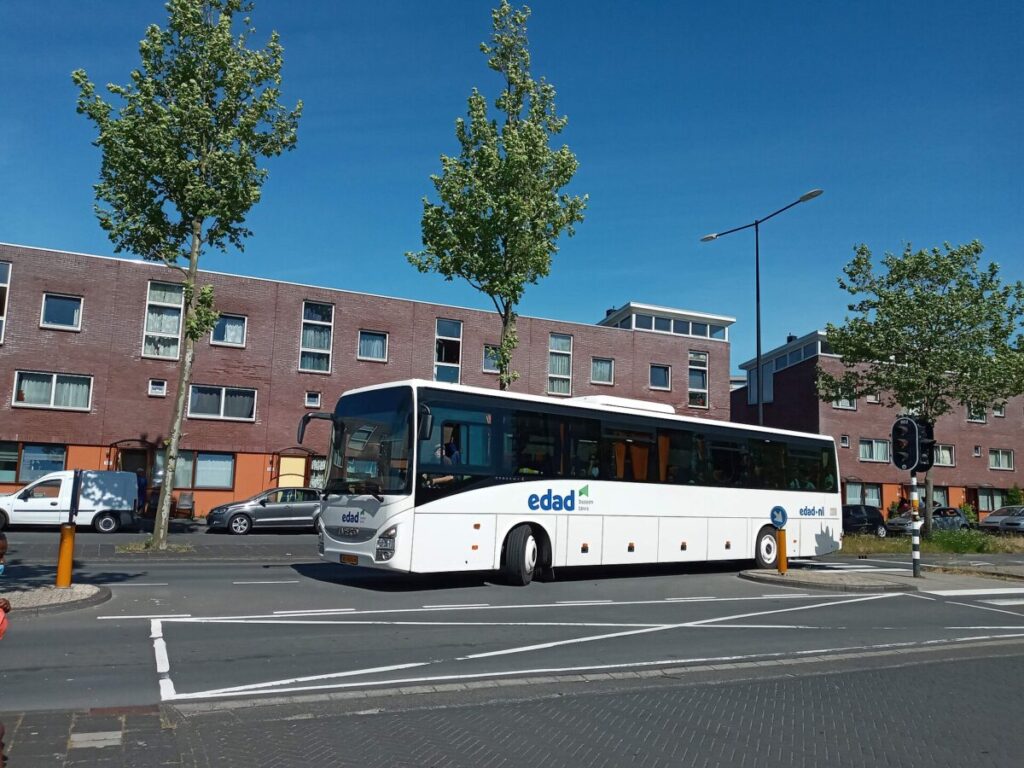 Modern urban bus on a sunny street near brick buildings and greenery.