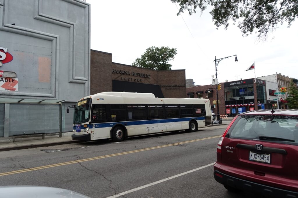 MTA bus at a busy urban street with shops, cars, and trees lining the road.