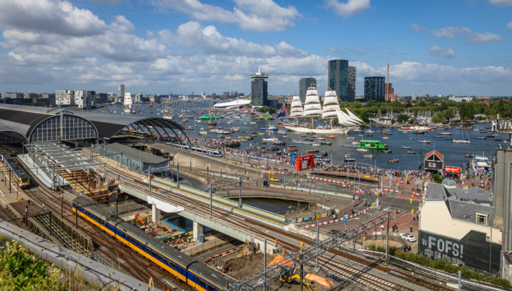 Vibrant Amsterdam waterfront scene with a sailing ship, crowd, and bustling Central Station.